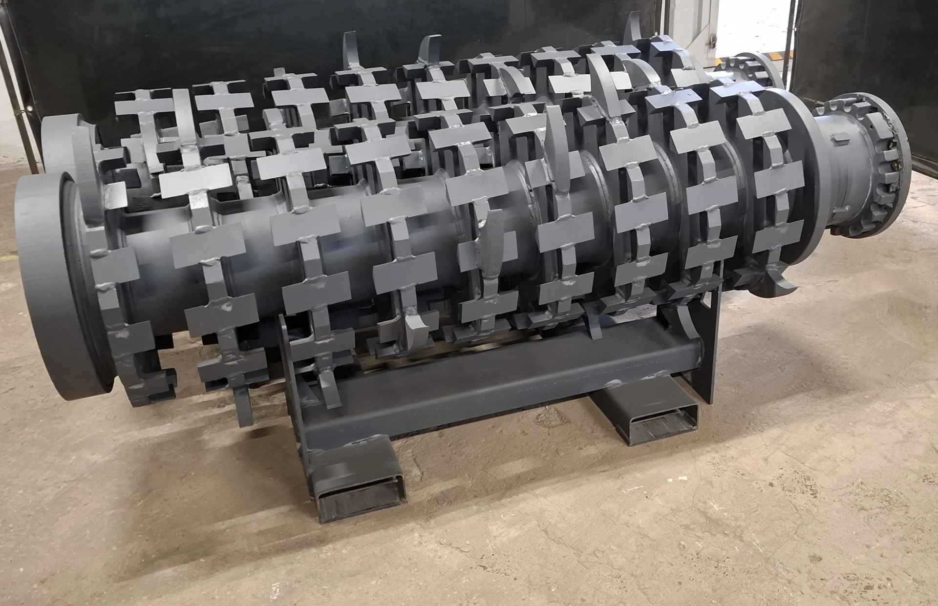A single, large gray industrial shredder T-shaft, featuring rows of durable T-shaped cutting elements, resting on a support frame.
